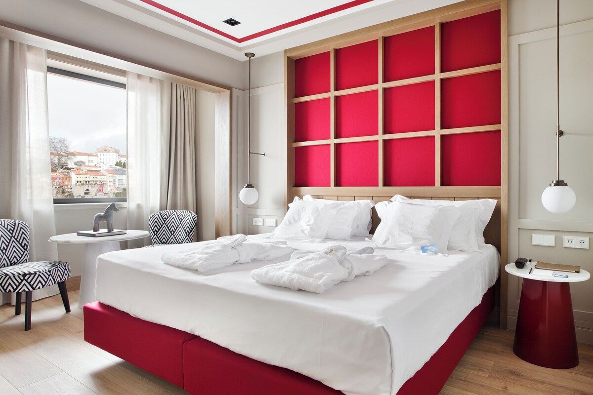 A modern hotel room with a large bed, a view of the city, a small round table, and a chair.