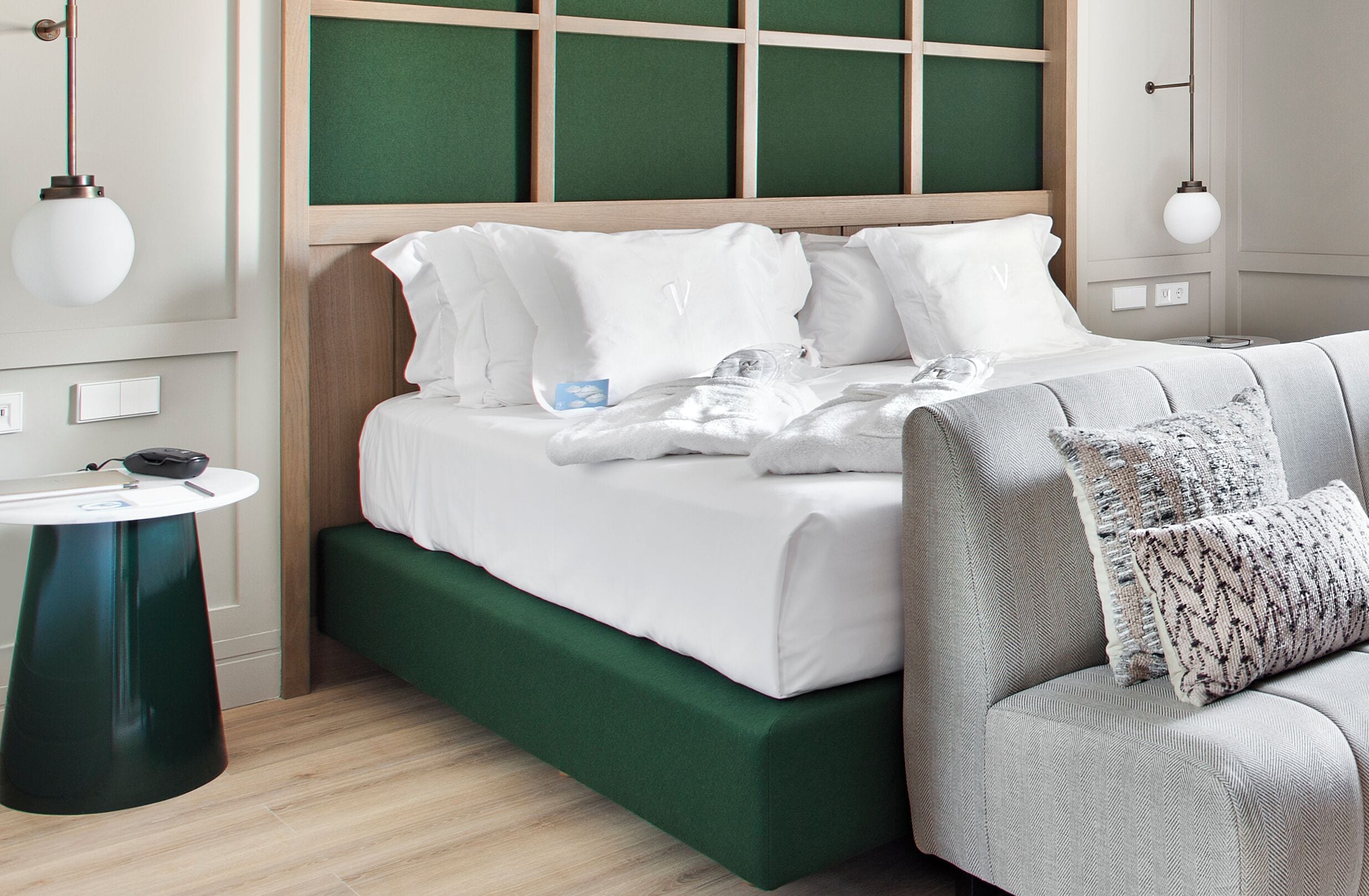 A neatly made bed with white linens, a green upholstered bench, and a round table with a black object on it.