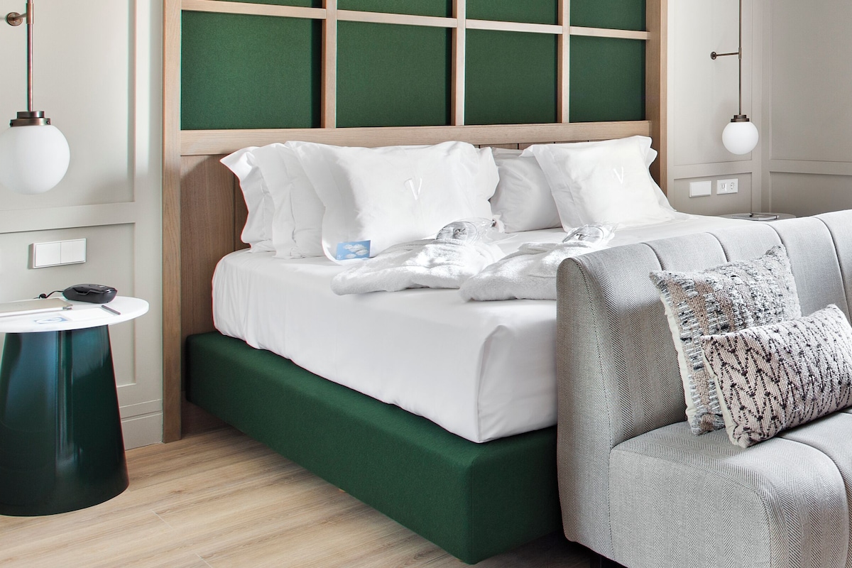 A neatly made bed with white linens, a green upholstered bench, and a round table with a black object on it.