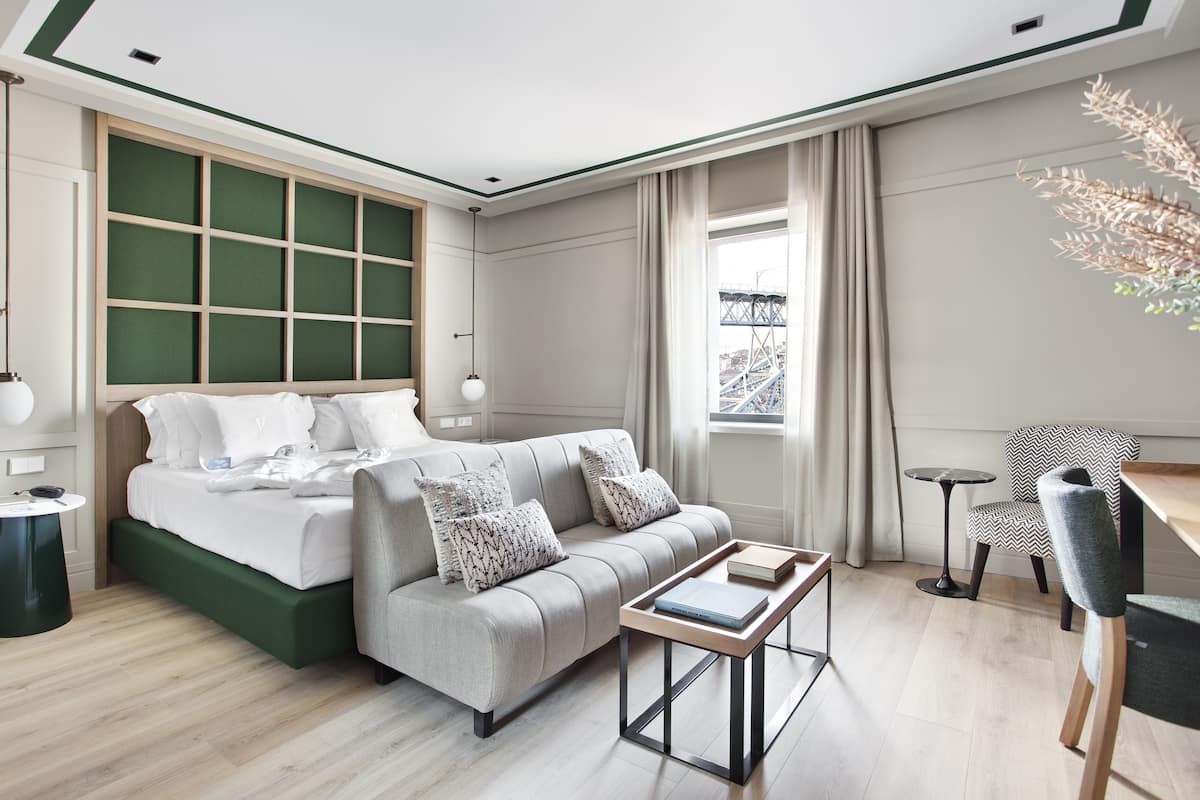 Een moderne hotelkamer met een bed, een bank en een klein tafeltje. Er is een raam met uitzicht op een brug.