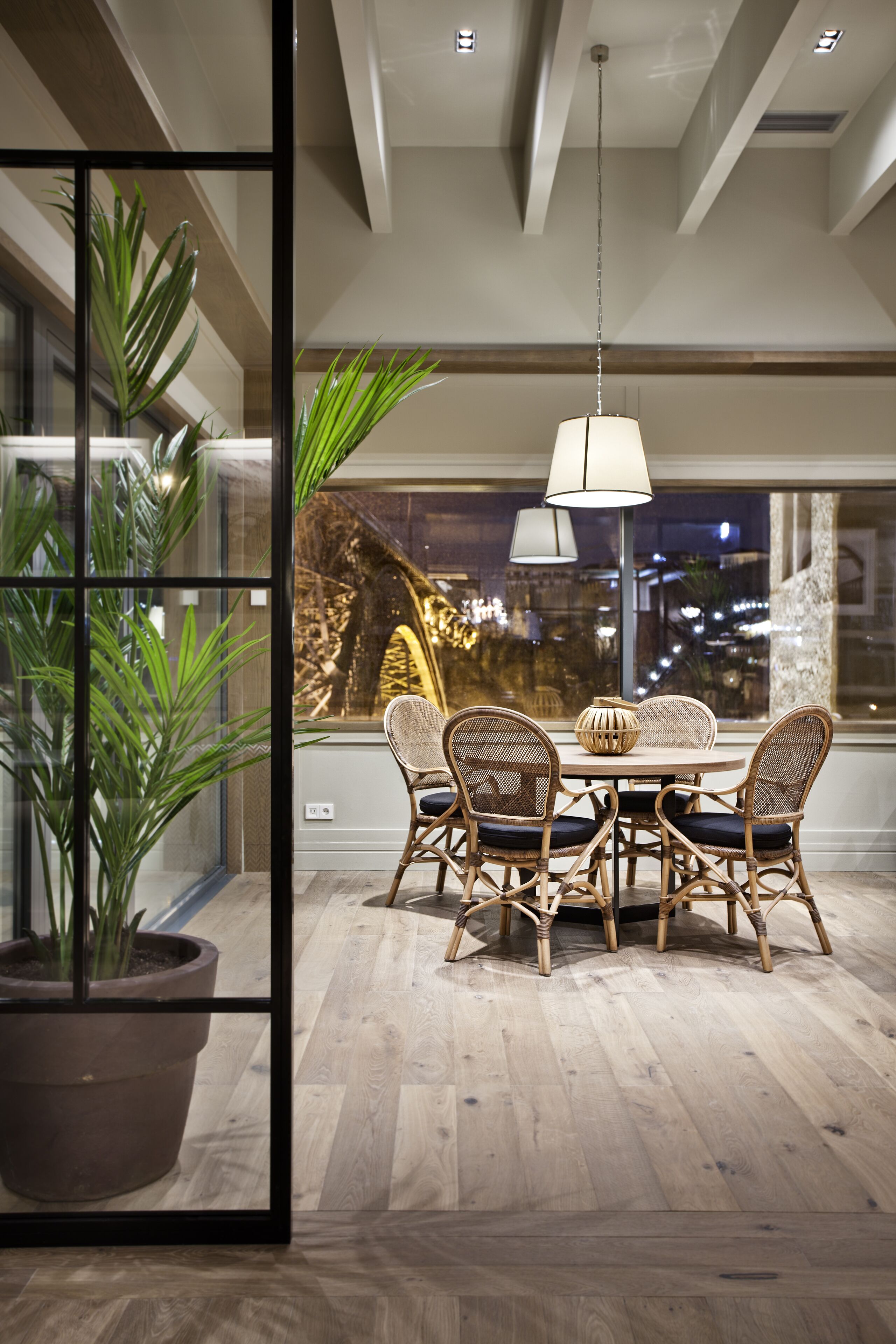 A dining area with a round table and wicker chairs, a hanging light fixture, and a view of the city through large windows.