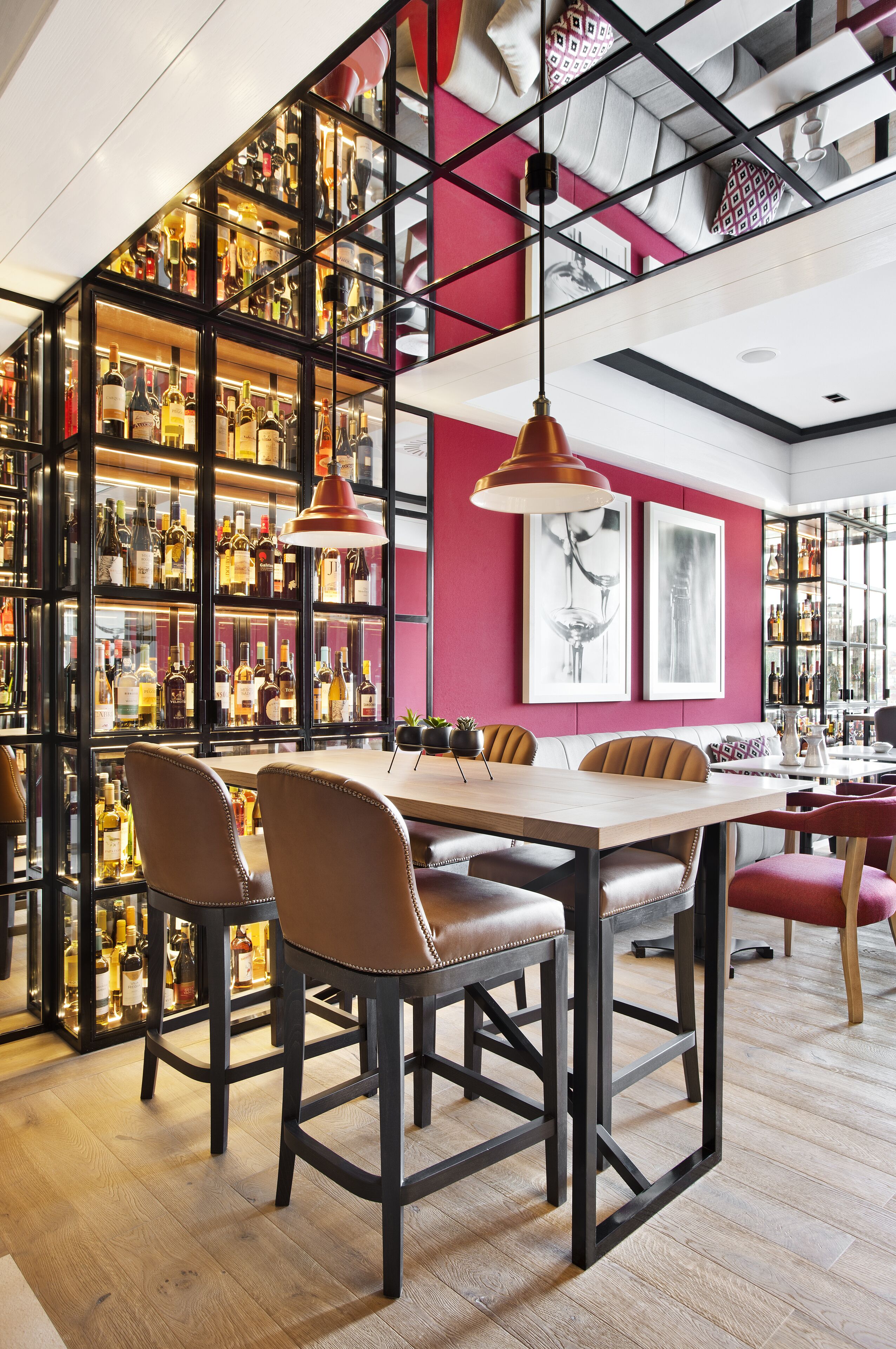 Een moderne bar met een lange houten tafel, comfortabele stoelen en een glazen vitrine vol flessen.