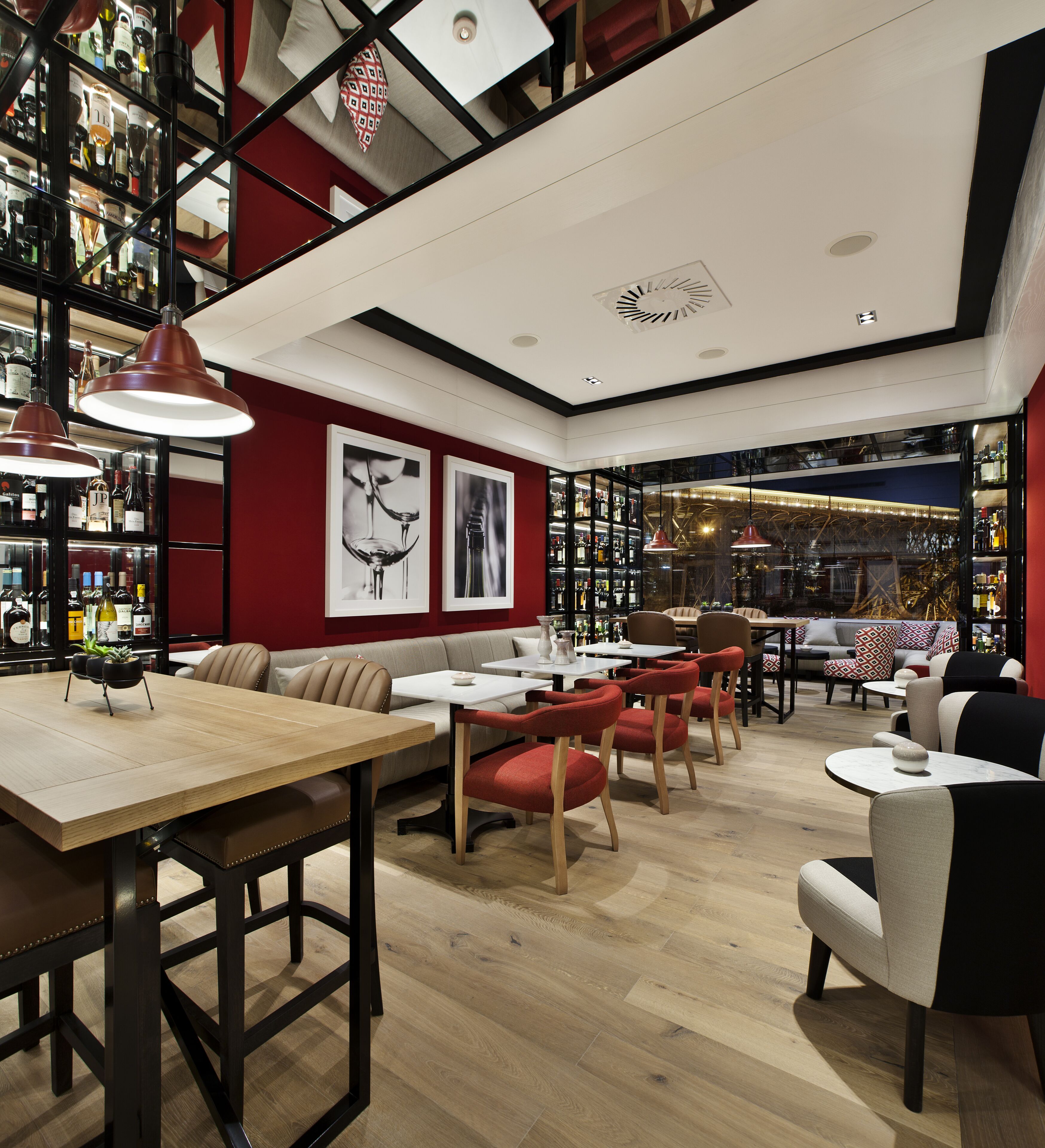 A modern bar with wooden tables and red chairs, a red wall with framed artwork, and a visible liquor shelf.
