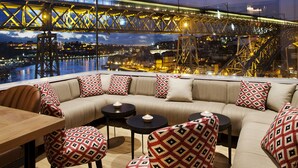 A modern lounge with a curved sofa, patterned armchairs, and a wooden table. A large window offers a view of a bridge and cityscape. - Hotel Vincci Ponte de Ferro (Vila Nova de Gaia)