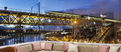 A modern lounge with a curved sofa, patterned armchairs, and a wooden table. A large window offers a view of a bridge and cityscape.