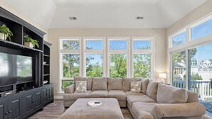 A living room with a sectional sofa, a flat-screen TV, dark cabinets, and large windows with a view of trees and houses. - It's All Good In Corolla: Soundside, Pool, Spa (Corolla)