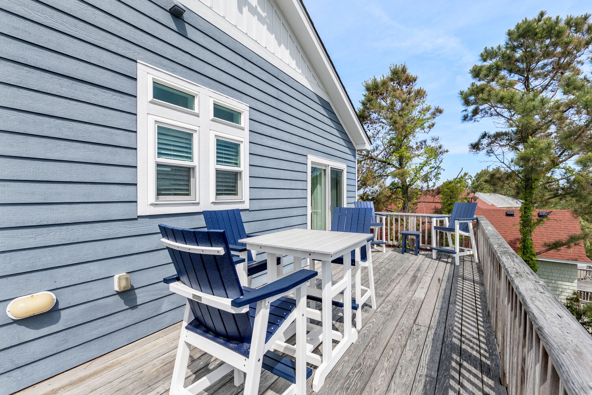 A wooden deck with blue Adirondack chairs and a white table, overlooking a residential area with pine trees and a house with a red roof.
