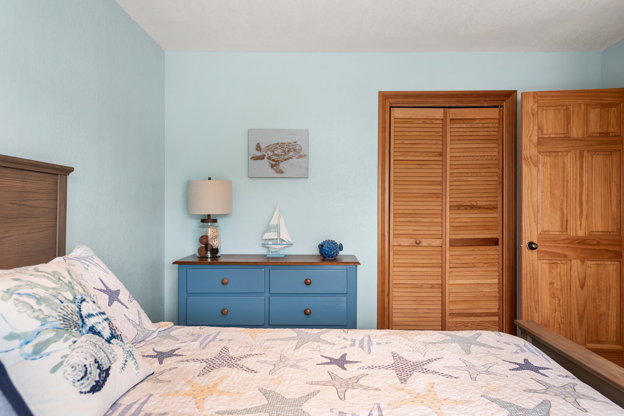 A bedroom with a bed, a dresser, a nightstand, a lamp, and a wooden wardrobe.