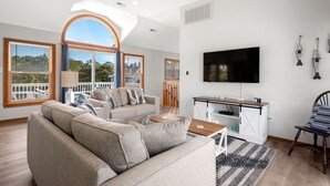Un vaste salon comprenant un grand canapé, une table basse et un téléviseur fixé au mur. Il y a également un balcon avec une balustrade et une vue sur l’extérieur.