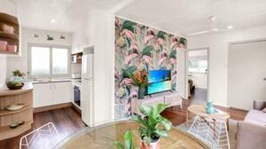 Una cocina moderna con gabinetes blancos, una zona de comedor con una mesa redonda de vidrio y una sala de estar con televisor y papel tapiz tropical.