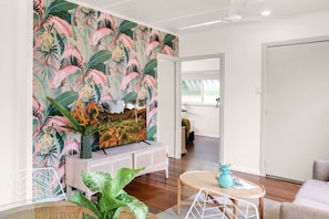 A living room with a tropical wallpaper, a flat-screen TV, a white console table, a round wooden coffee table, and a large green plant. - Stylish & Pet-Friendly Getaway – Your Tropical Escape Awaits (Parramatta Park)