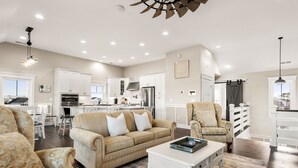 Amplia sala de estar con sofá, sillones y una mesa de centro. Se observa una cocina con armarios blancos y un comedor con mesa y sillas.