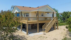Ein Strandhaus mit zwei Etagen, einer Holzterrasse und einer Treppe, die zum oberen Stockwerk führt.