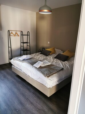 Ein ordentlich bezogenes Bett mit grauer Decke und Kissen, ein Wäscheständer aus Holz und eine hängende Lampenleuchte.
