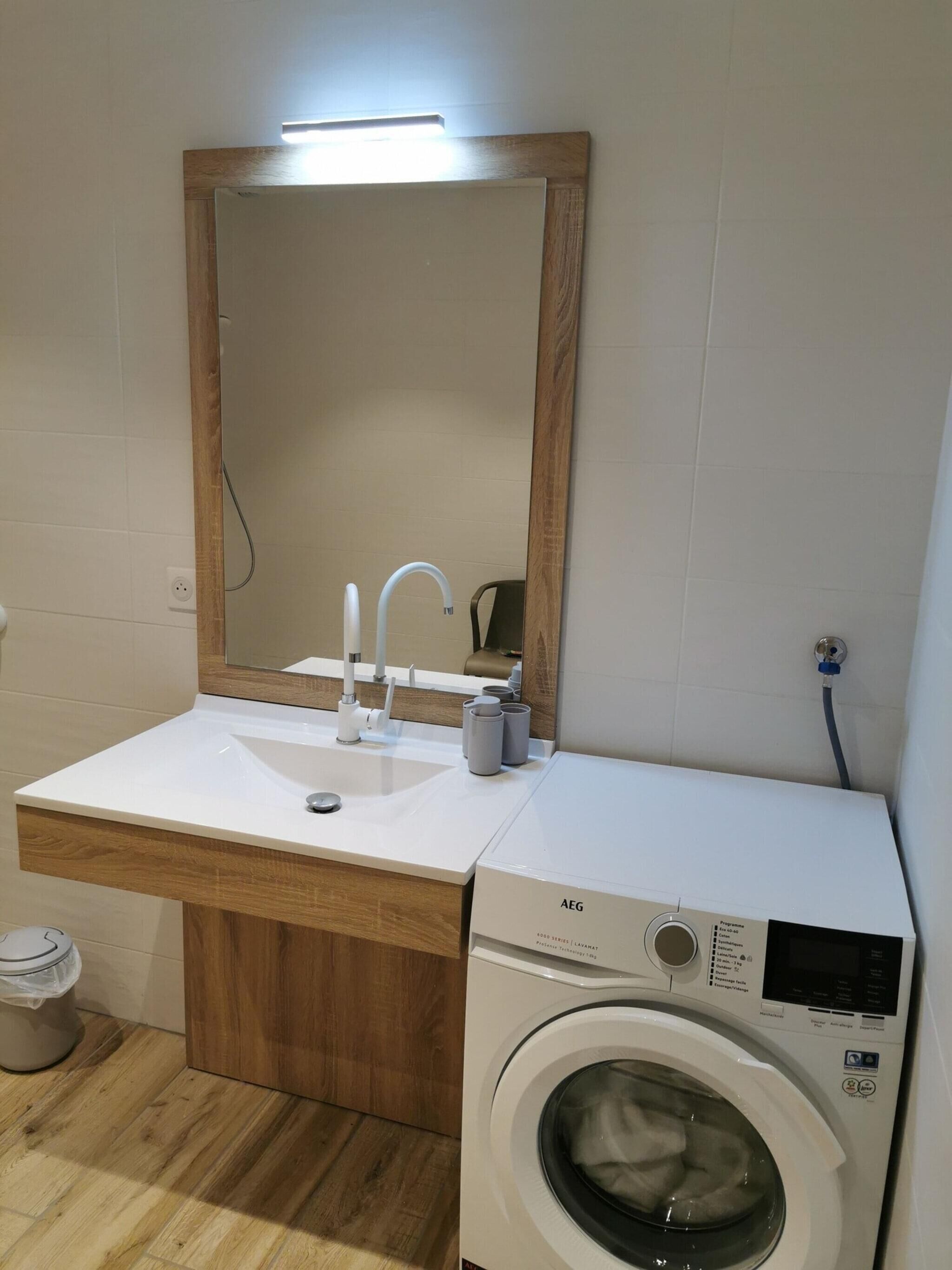 A compact bathroom with a wooden vanity, a sink, a mirror, and a front-loading washing machine.