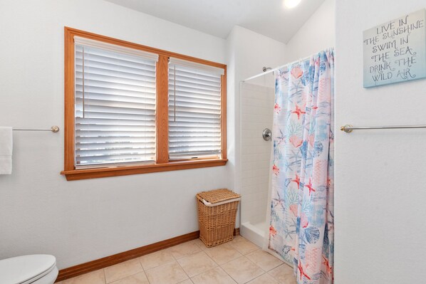 A bathroom with a window, a shower curtain with a sea theme, and a wicker basket. - Livin': Oceanfront, Pool, Spa, Elevator (Corolla)