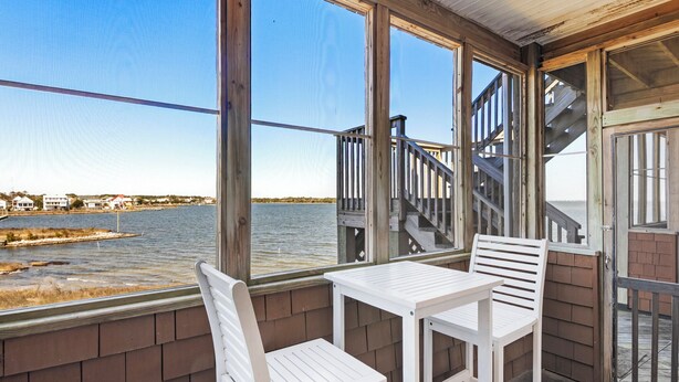 A balcony with white chairs and a table, overlooking a body of water and a dock. - North Bay Retreat: Waterfront, Shared Pool, Fitness Center, Short Stays Anytime (Manteo)