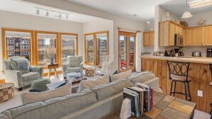 A spacious living room with a large sofa, two armchairs, a coffee table, and a kitchen area with wooden cabinets and a bar stool. - North Bay Retreat: Waterfront, Shared Pool, Fitness Center, Short Stays Anytime (Manteo)
