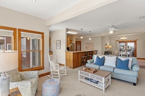 A spacious living room with a sofa, ottoman, and rocking chair, a kitchen area with a bar, and a dining area with a chandelier. - North Bay Retreat: Waterfront, Shared Pool, Fitness Center, Short Stays Anytime (Manteo)