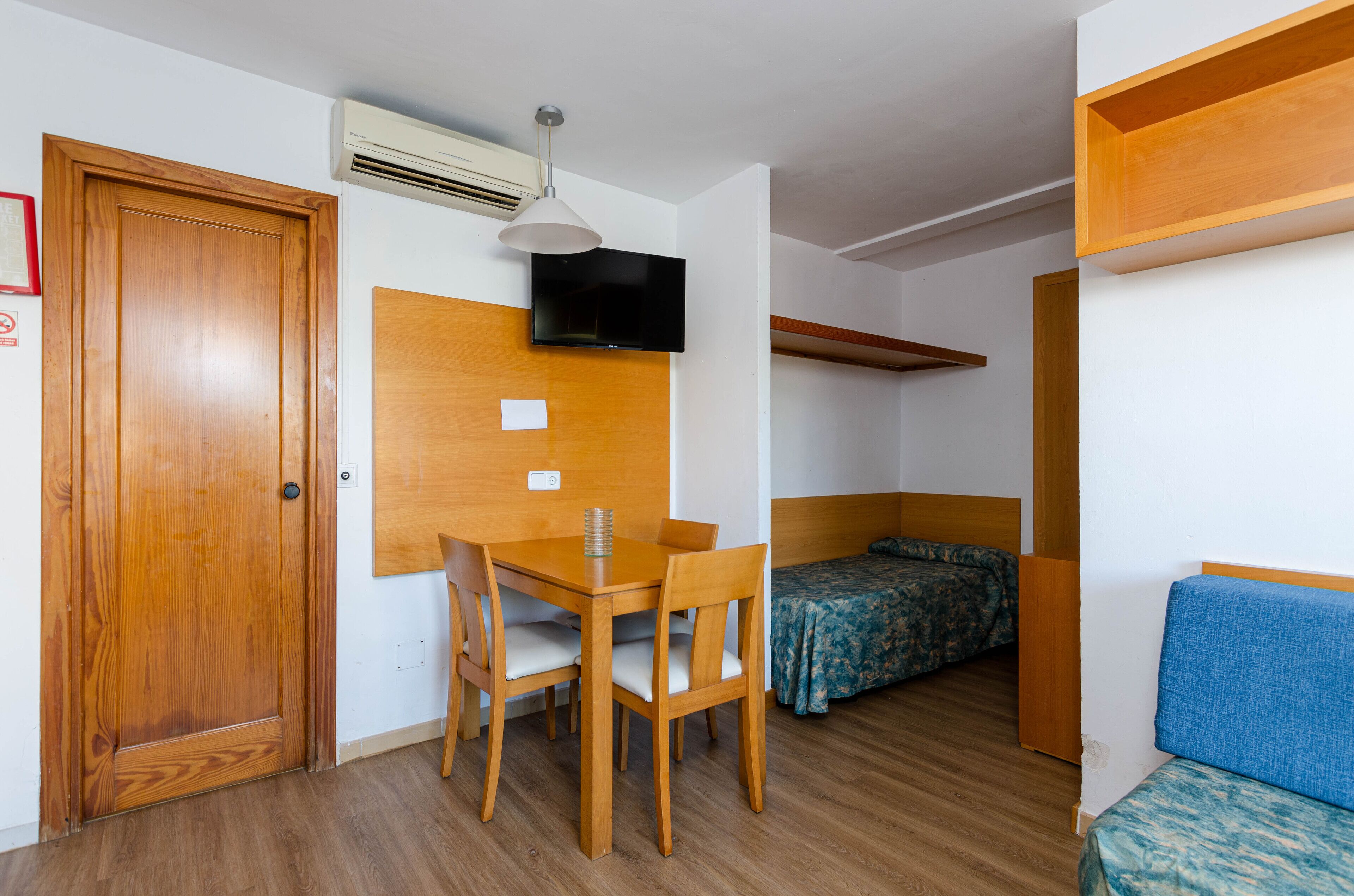 Una habitación pequeña con puerta de madera, una mesa con sillas, una cama con colcha estampada y un televisor montado en la pared.