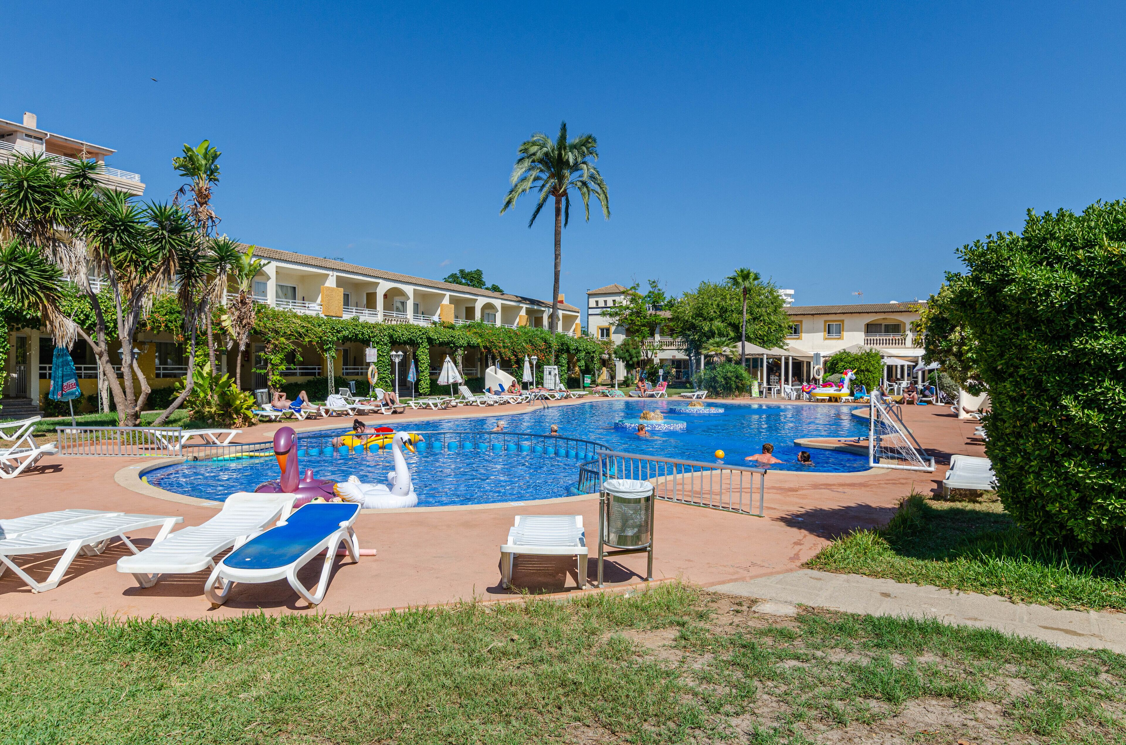 Ein Hotelpoolbereich mit mehreren Liegestühlen, einem zentralen Schwimmbecken und Palmen.