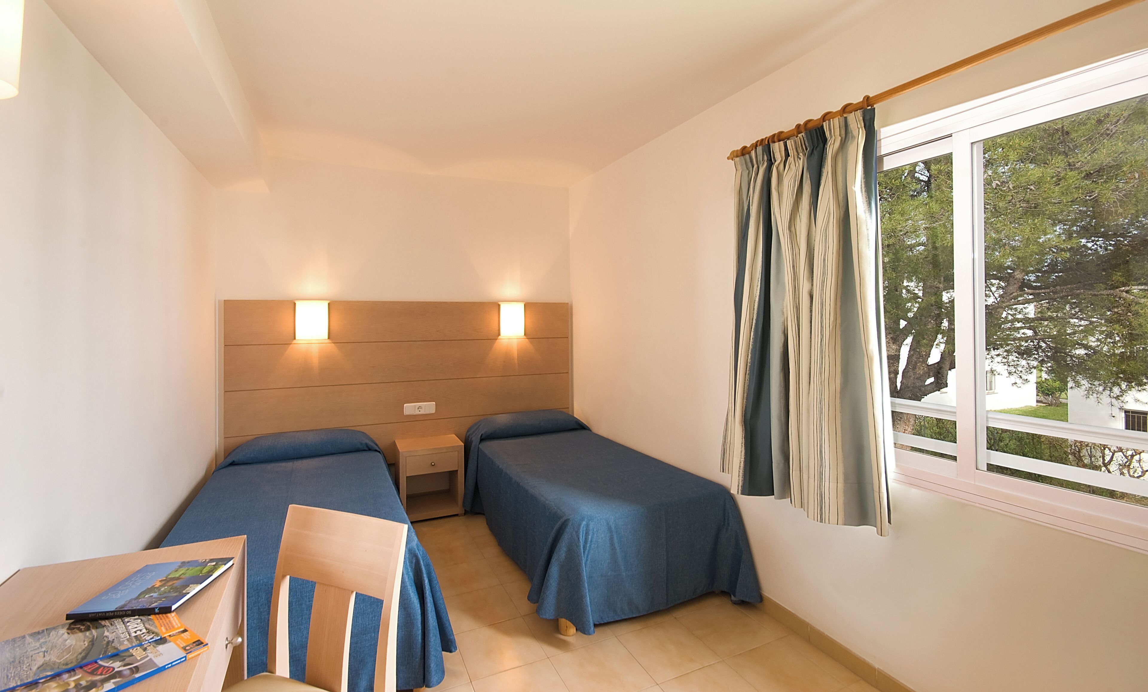 A small, well-lit room with two single beds, a desk with a chair, and a window with curtains.