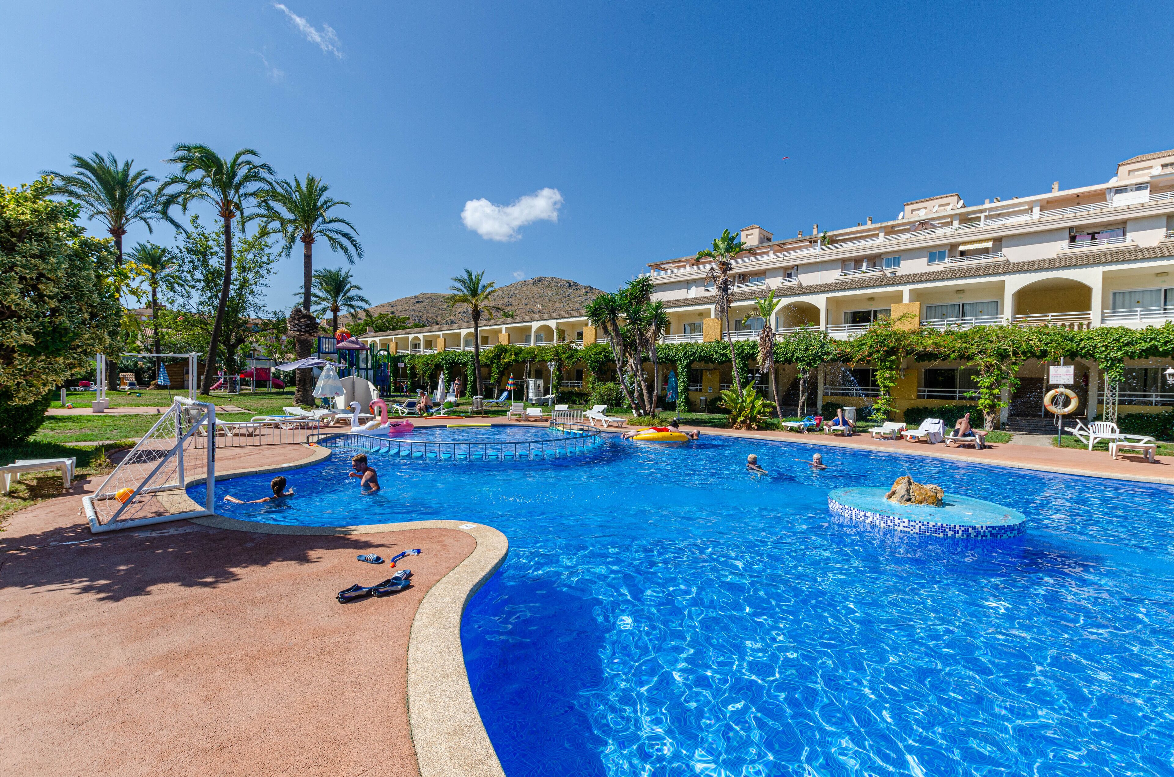 A large swimming pool with a diving board and lounge chairs, surrounded by palm trees and a building with multiple balconies.