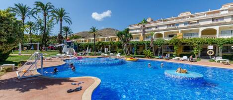 A large swimming pool with a diving board and lounge chairs, surrounded by palm trees and a building with multiple balconies.