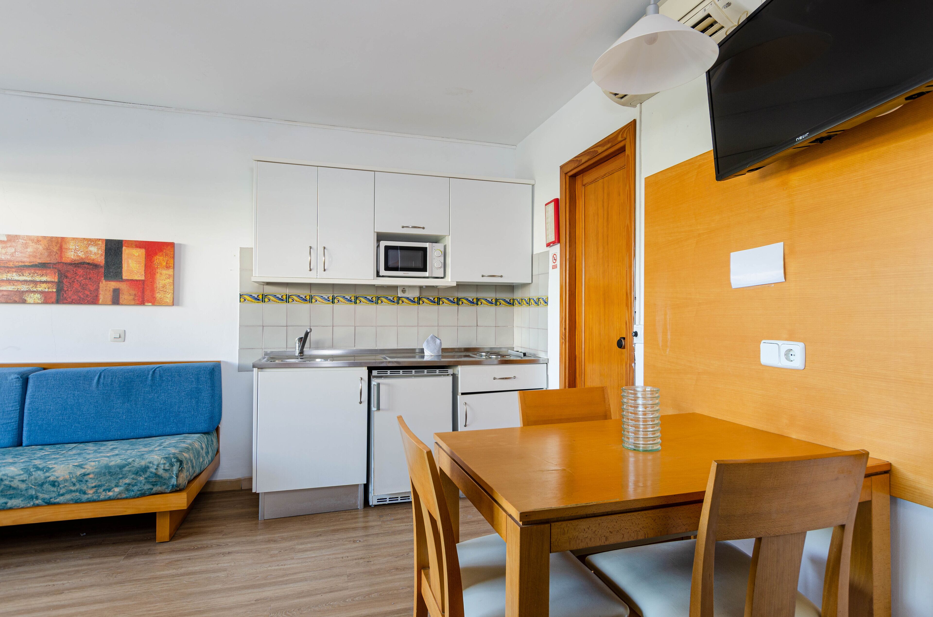 Cocina compacta con armarios blancos, un microondas y un refrigerador pequeño. Hay una mesa de comedor de madera con sillas y un sofá azul en la sala.