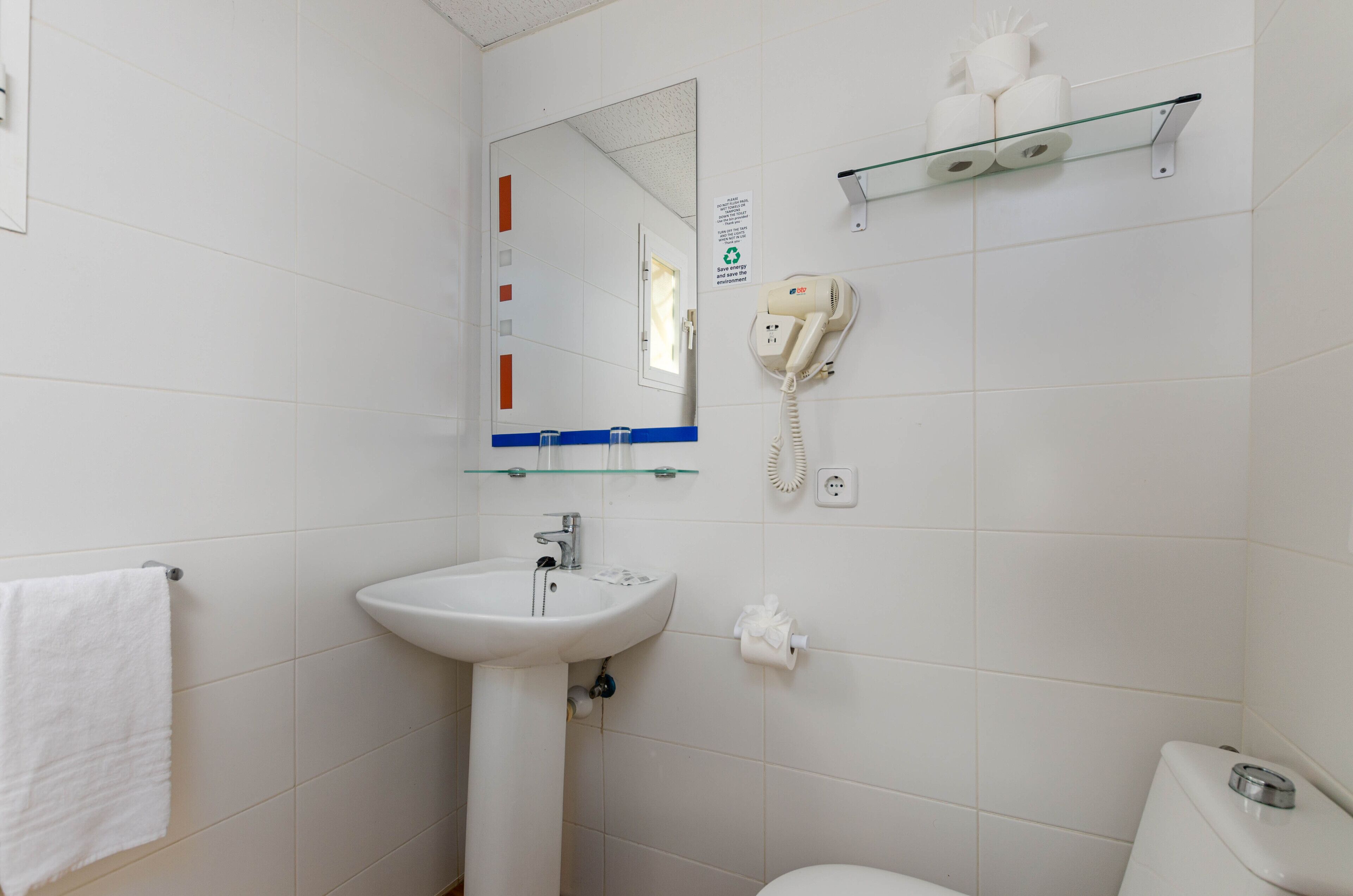 Un baño con lavamanos blanco, espejo, inodoro y estante de vidrio con artículos de tocador.
