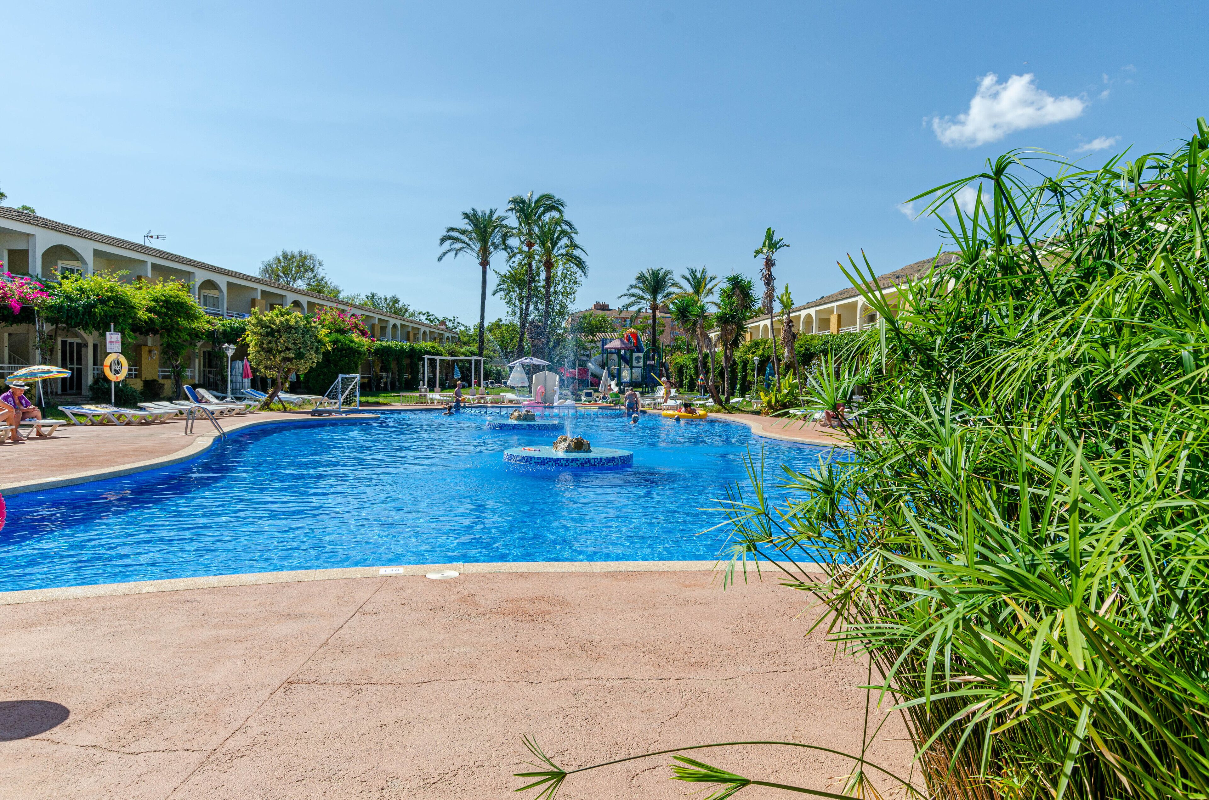 A swimming pool with fountains, surrounded by palm trees and a hotel building.