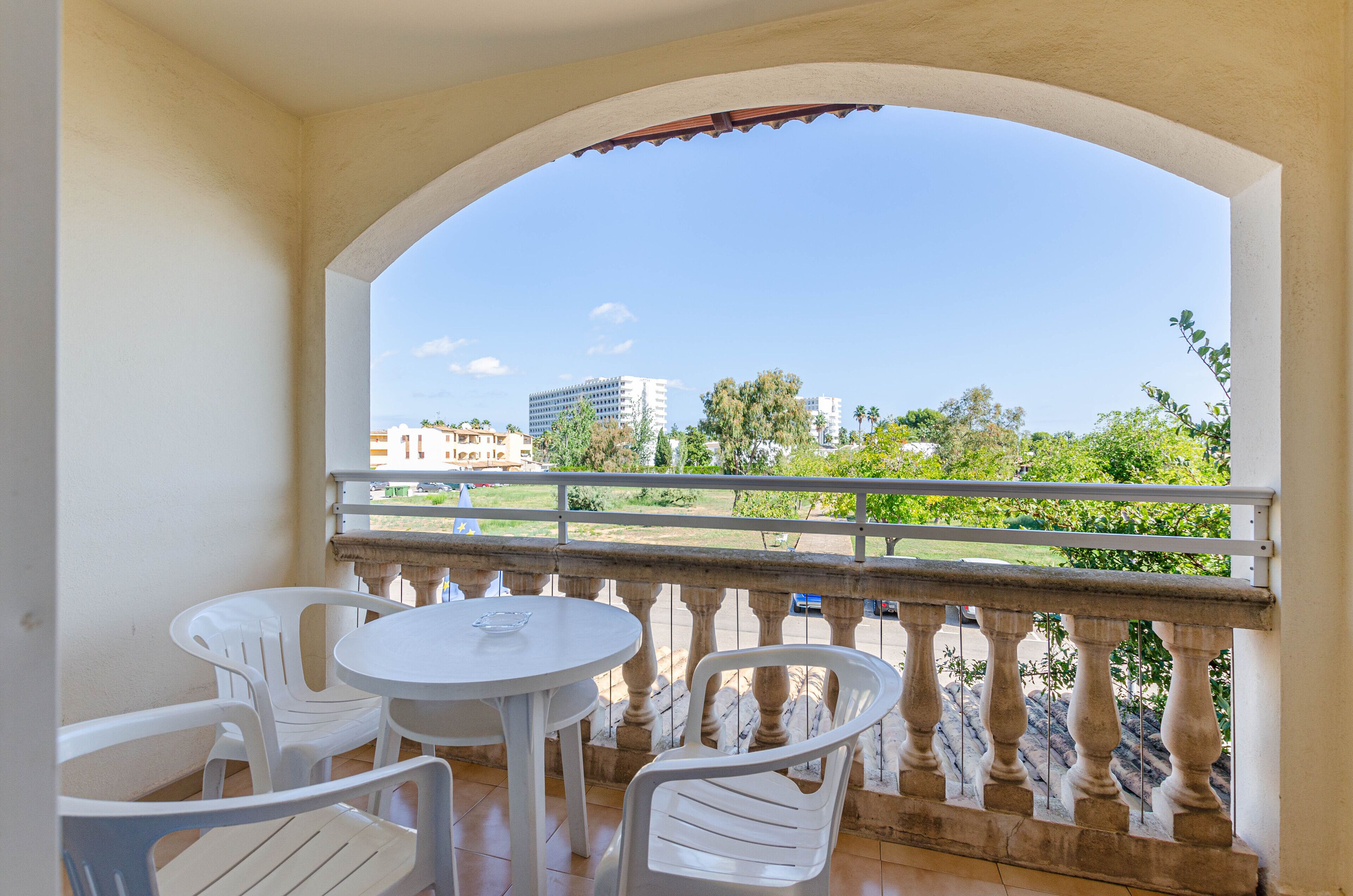 A balcony with white plastic chairs and a table, overlooking a green landscape and buildings.