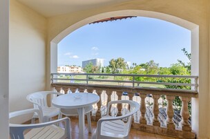 A balcony with white plastic chairs and a table, overlooking a green landscape and buildings.