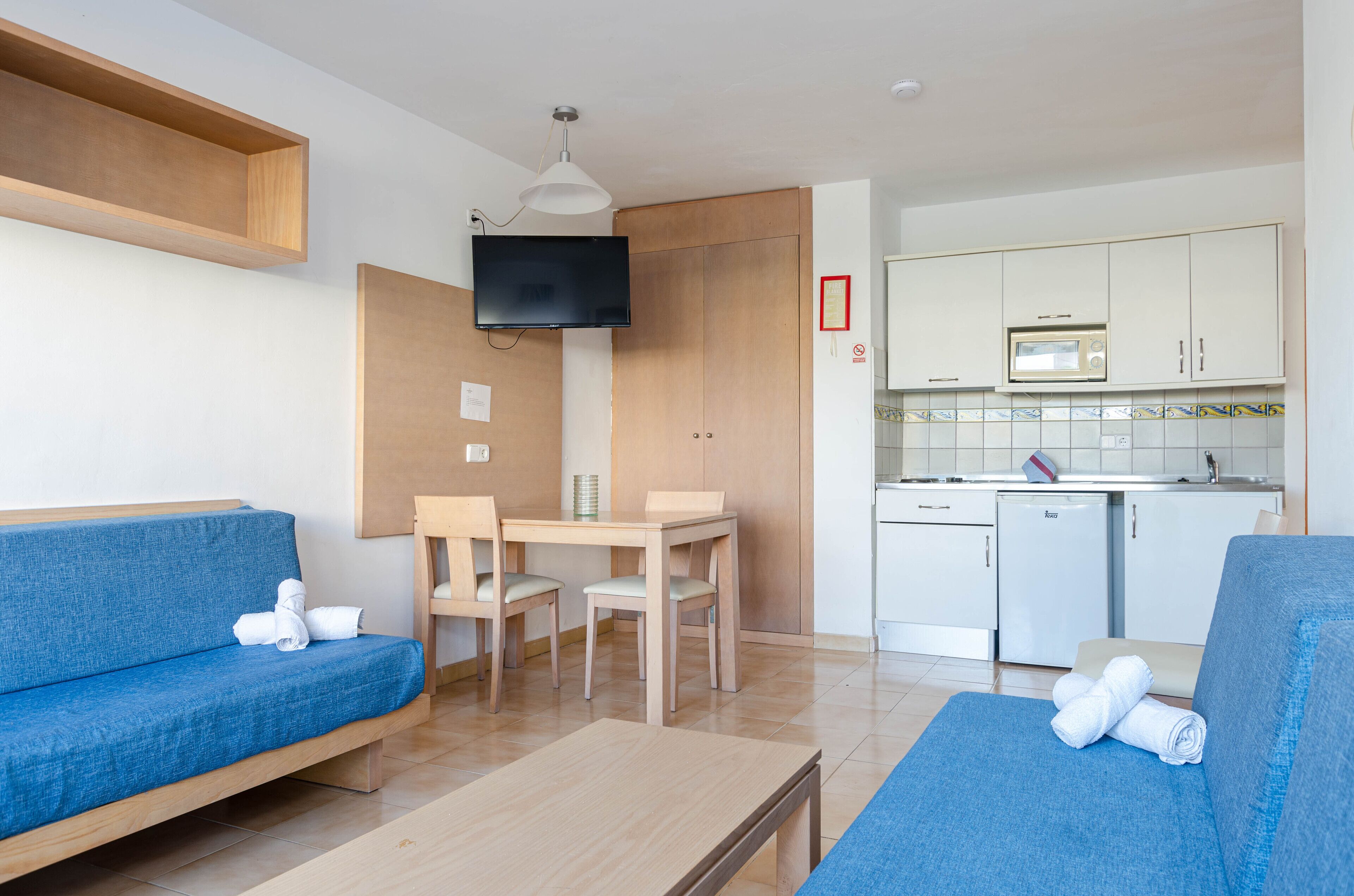 Un salon moderne avec un canapé bleu, une table à manger en bois accompagnée de deux chaises, un téléviseur à écran plat et un coin cuisine équipé d’un four à micro-ondes et d’un réfrigérateur.