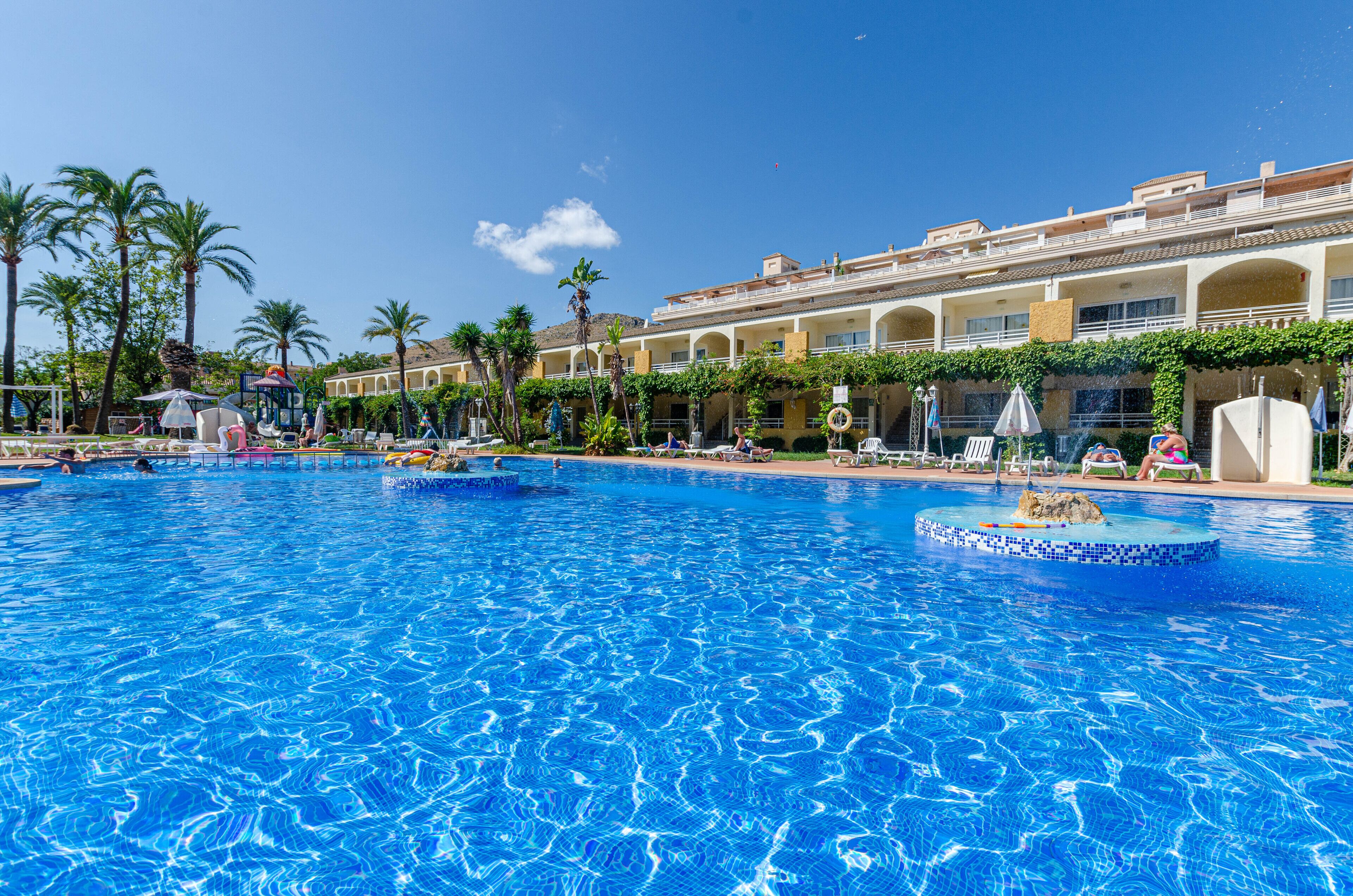 A large outdoor swimming pool with multiple sections, surrounded by palm trees and a hotel building with balconies.