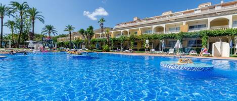 A large outdoor swimming pool with multiple sections, surrounded by palm trees and a hotel building with balconies.