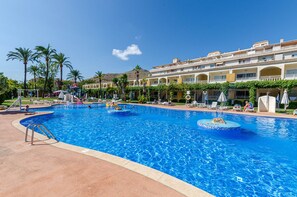 A large outdoor swimming pool with multiple lanes, surrounded by lounge chairs and umbrellas, and a building with balconies in the background. - Mariner Club Apartments (Alcúdia)