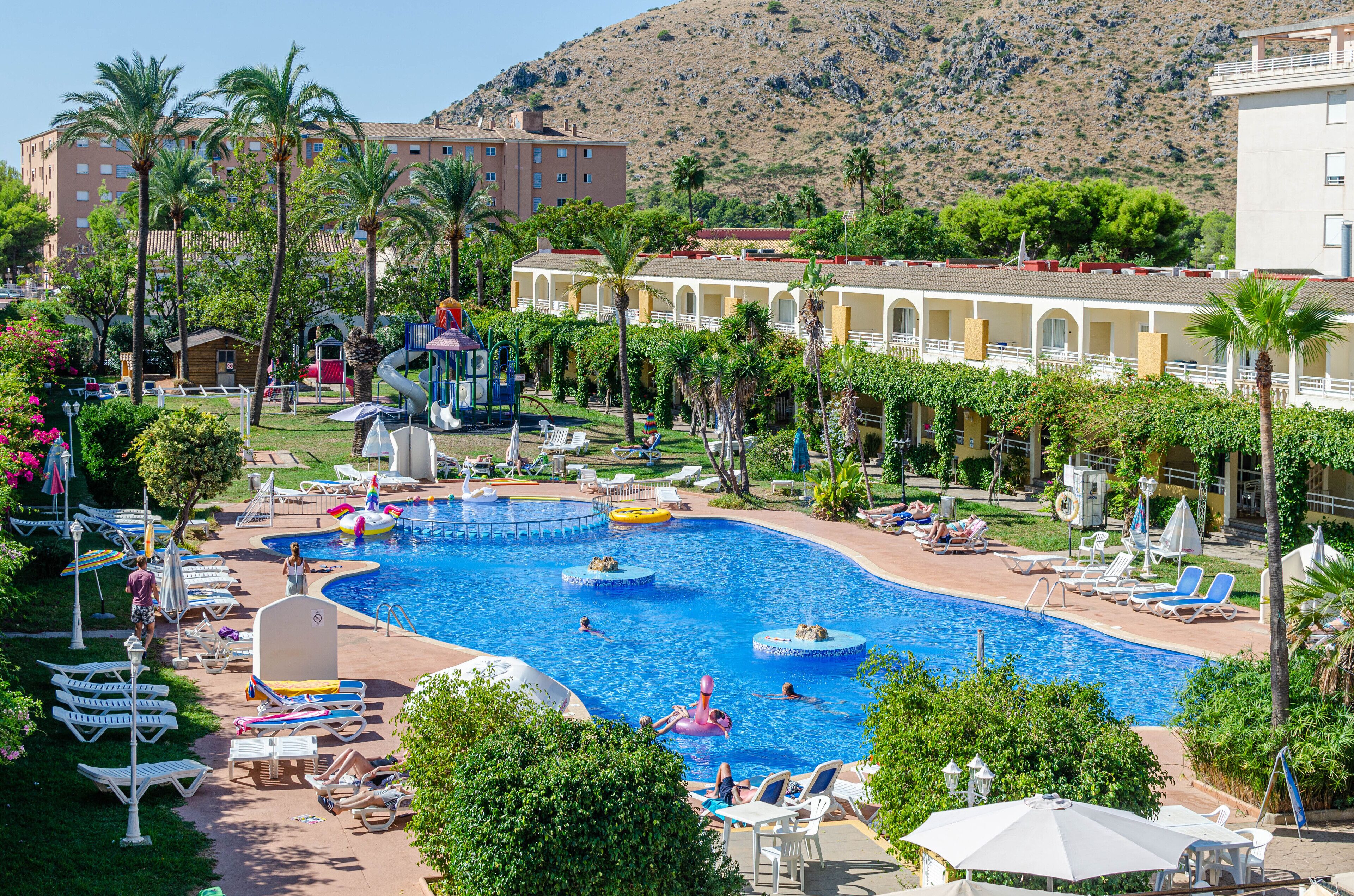 Un resort con una amplia piscina, tumbonas, palmeras y montañas al fondo.