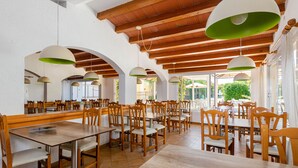 A spacious dining area with wooden tables and chairs, large pendant lights, and a view of the outdoors. - Mariner Club Apartments (Alcúdia)