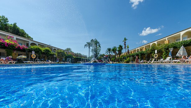 A clear blue pool with lounge chairs and umbrellas on both sides, surrounded by buildings with balconies and greenery. - Mariner Club Apartments (Alcúdia)