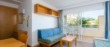 A room with a wooden desk, a sofa, a small table, and a balcony with a view of buildings and greenery.