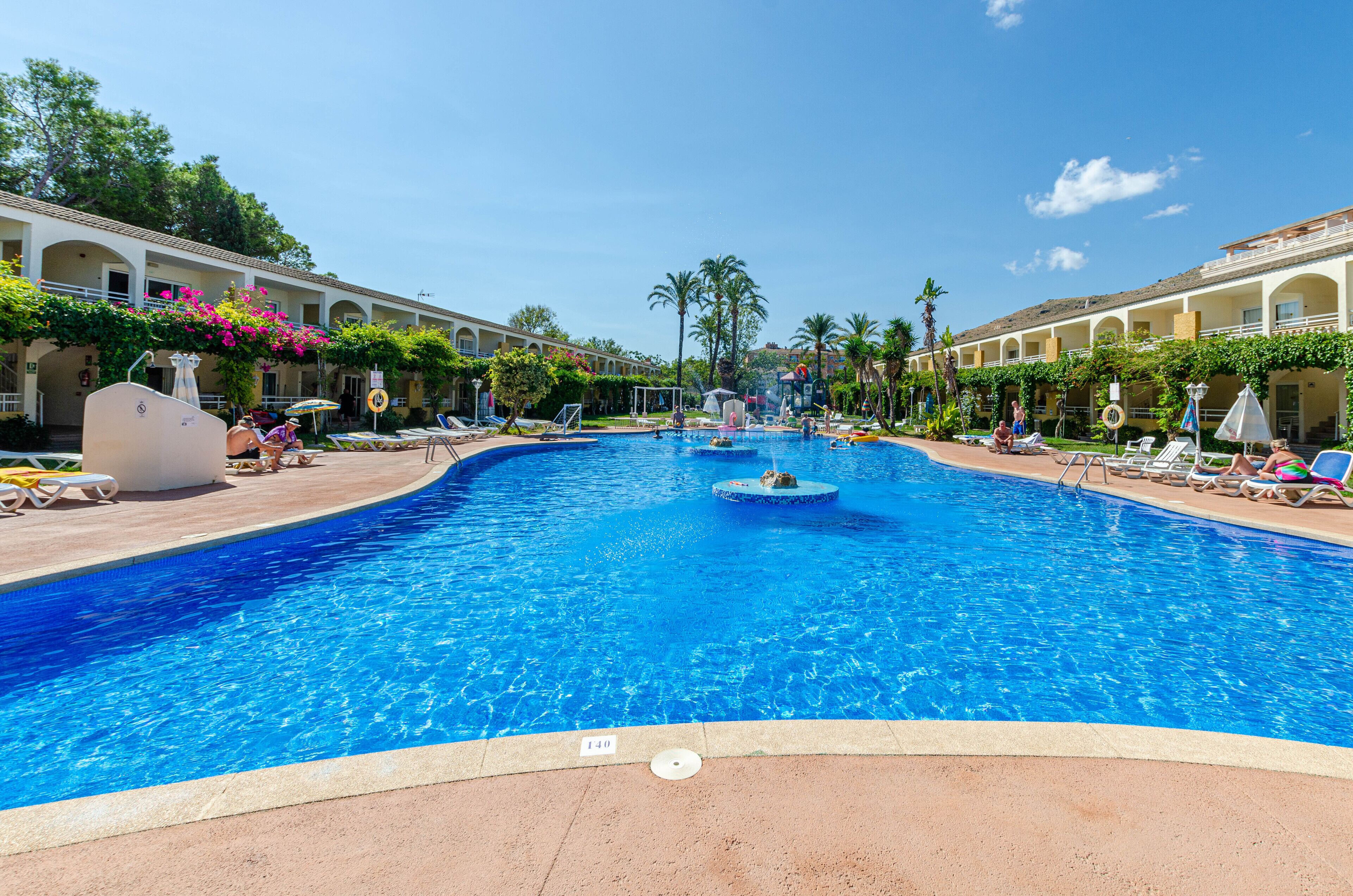 A large swimming pool with multiple lanes, surrounded by lounge chairs and umbrellas, and a building with a covered walkway.