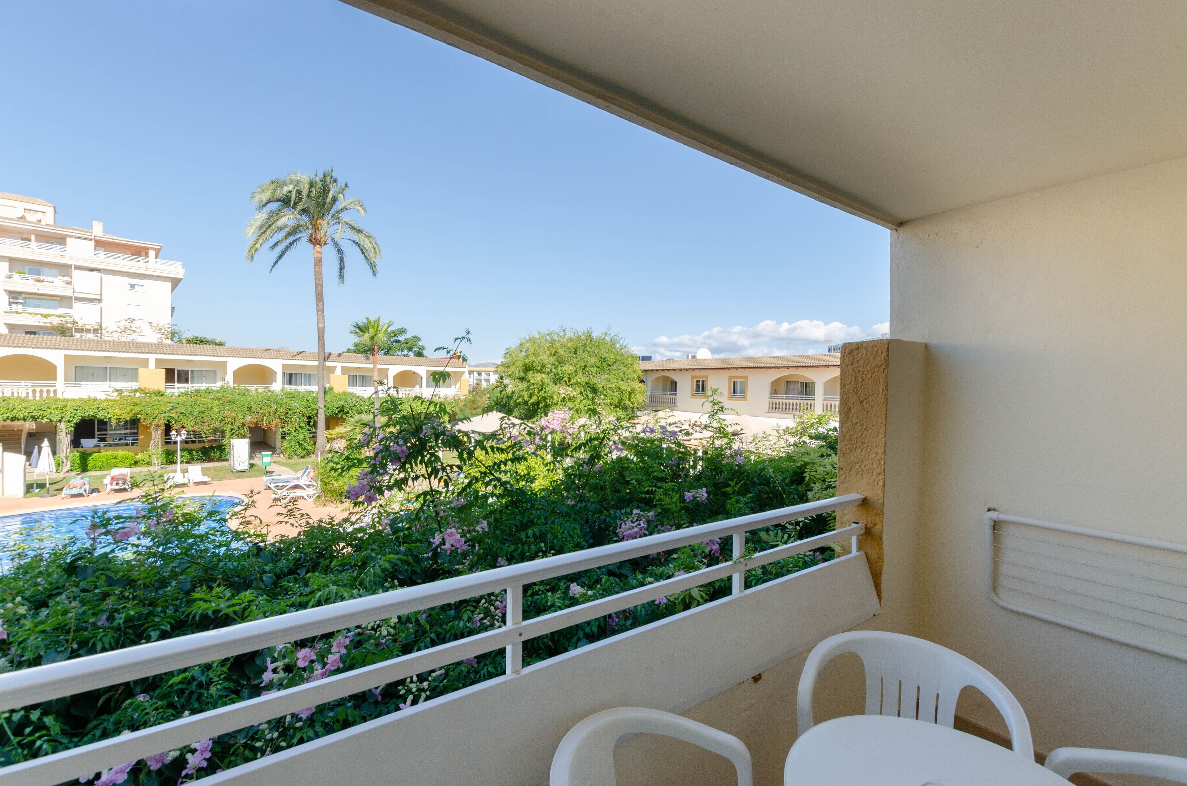 A balcony with white chairs and a table, overlooking a pool and lush greenery.
