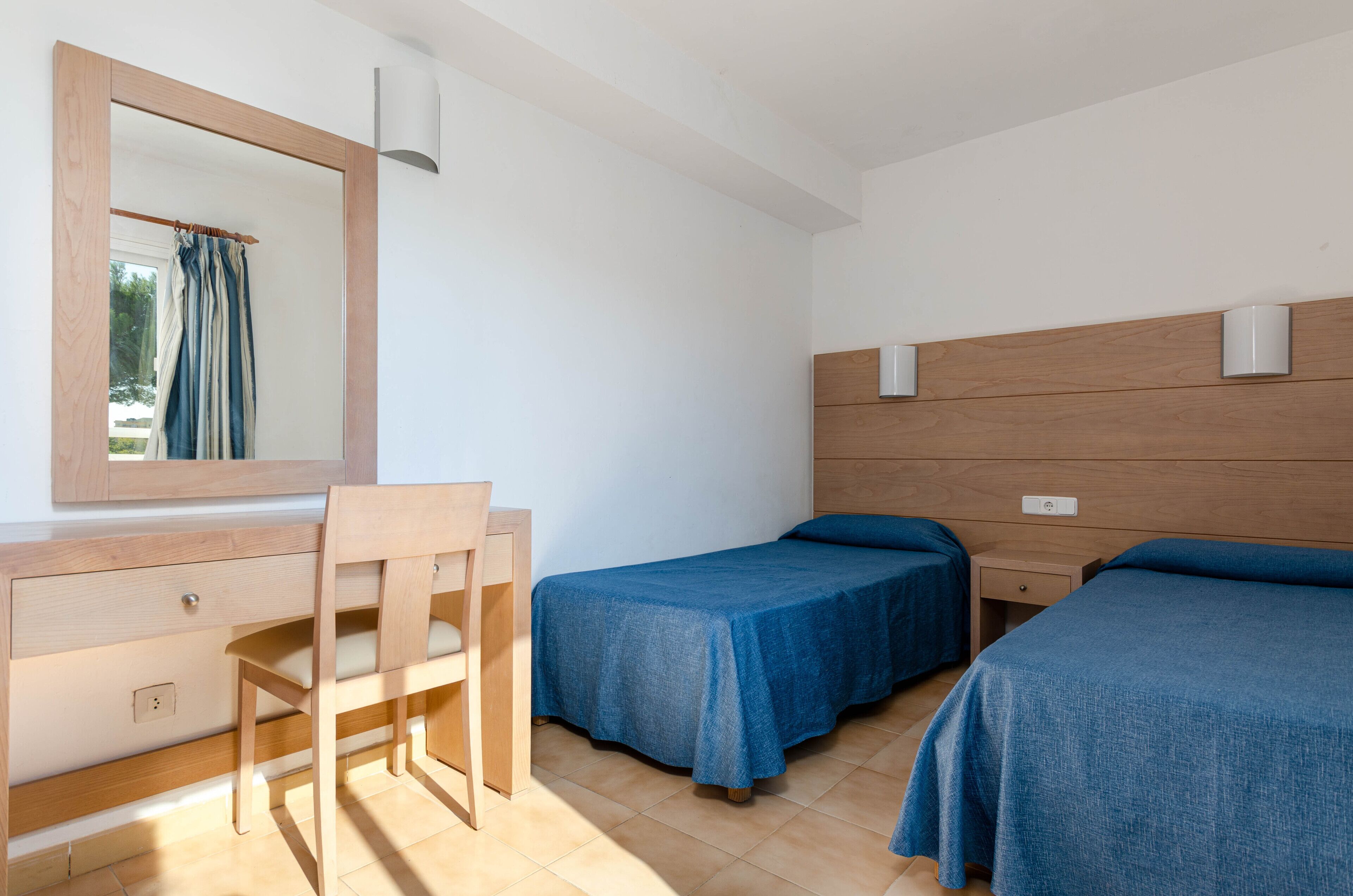 Een slaapkamer met een houten bureau, twee eenpersoonsbedden, een spiegel en een raam met gordijnen.
