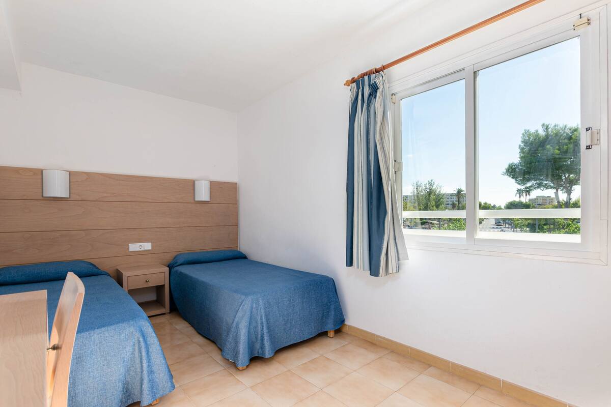 Ein Zimmer mit zwei Einzelbetten, einem Holzkopfteil, einem kleinen Nachttisch mit Schublade und einem Fenster mit Vorhang.