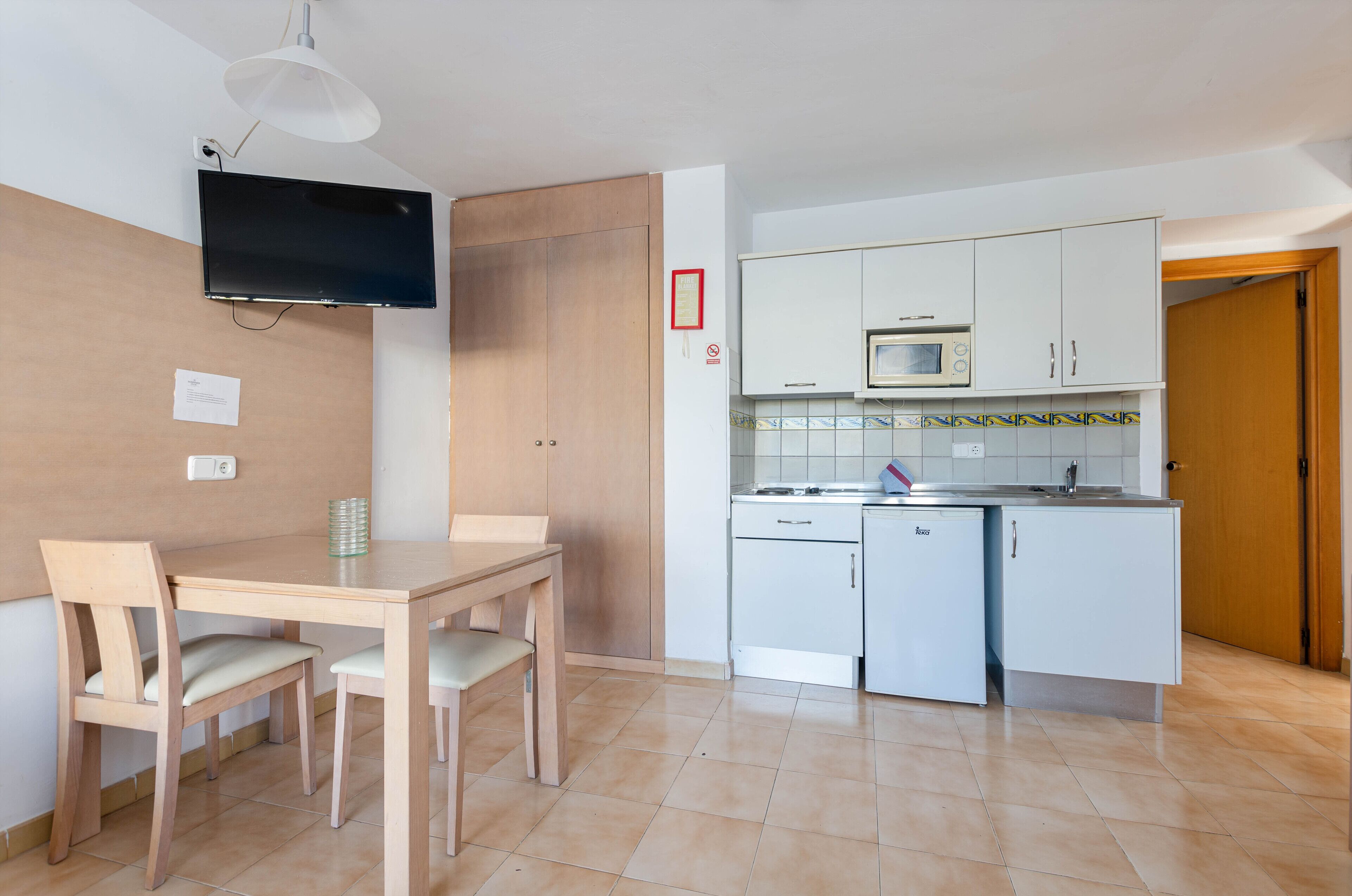 A compact kitchen with white cabinets, a microwave, and a small refrigerator. There is a wooden dining table with two chairs.