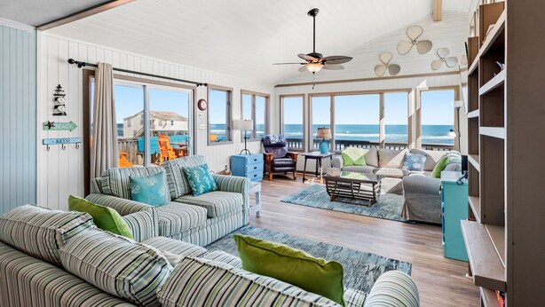 A spacious living room with a view of the ocean, featuring striped sofas, a ceiling fan, and a bookshelf.