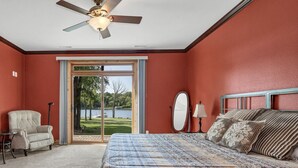 A bedroom with a large bed, a ceiling fan, a chair, a lamp, and a view of a lake through a glass door.