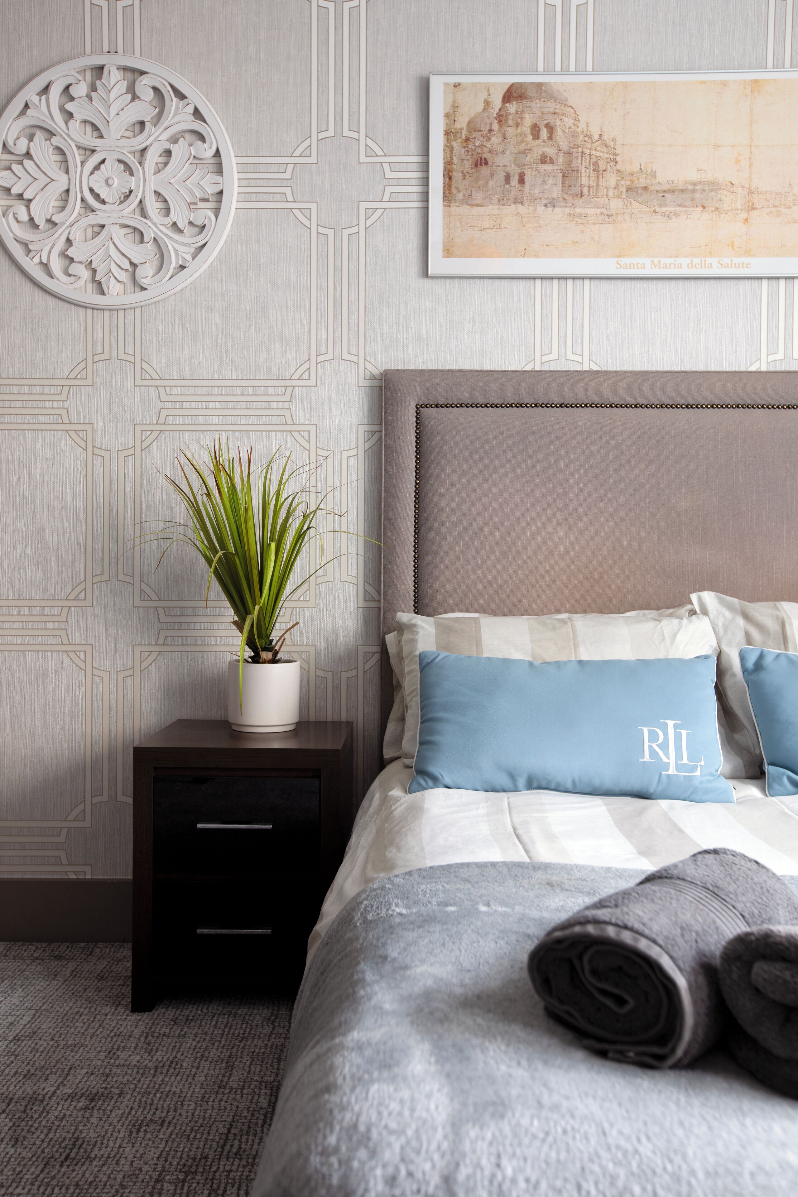 Un dormitorio con cama, almohadas, mesita de noche, planta en maceta y pared con diseño geométrico y un cuadro enmarcado.