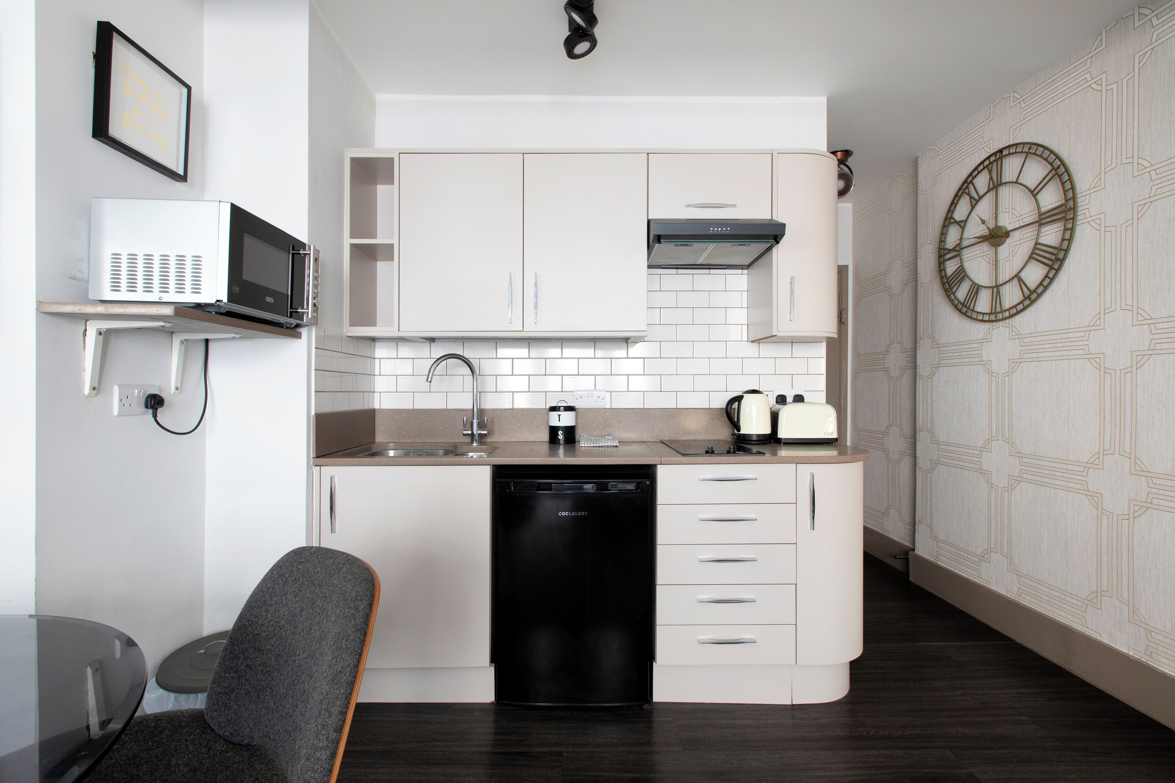 A compact kitchen with white cabinets, a dishwasher, a microwave, a toaster, and a clock on the wall.